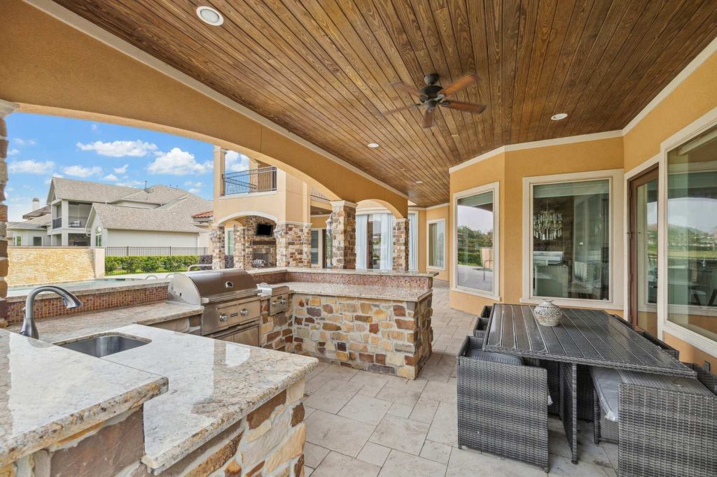 Spacious outdoor kitchen with granite counters, built-in grill, wicker dining set, and wooden ceiling overlooking scenic yard, handled by Real Estate Brokers Sugar Land.