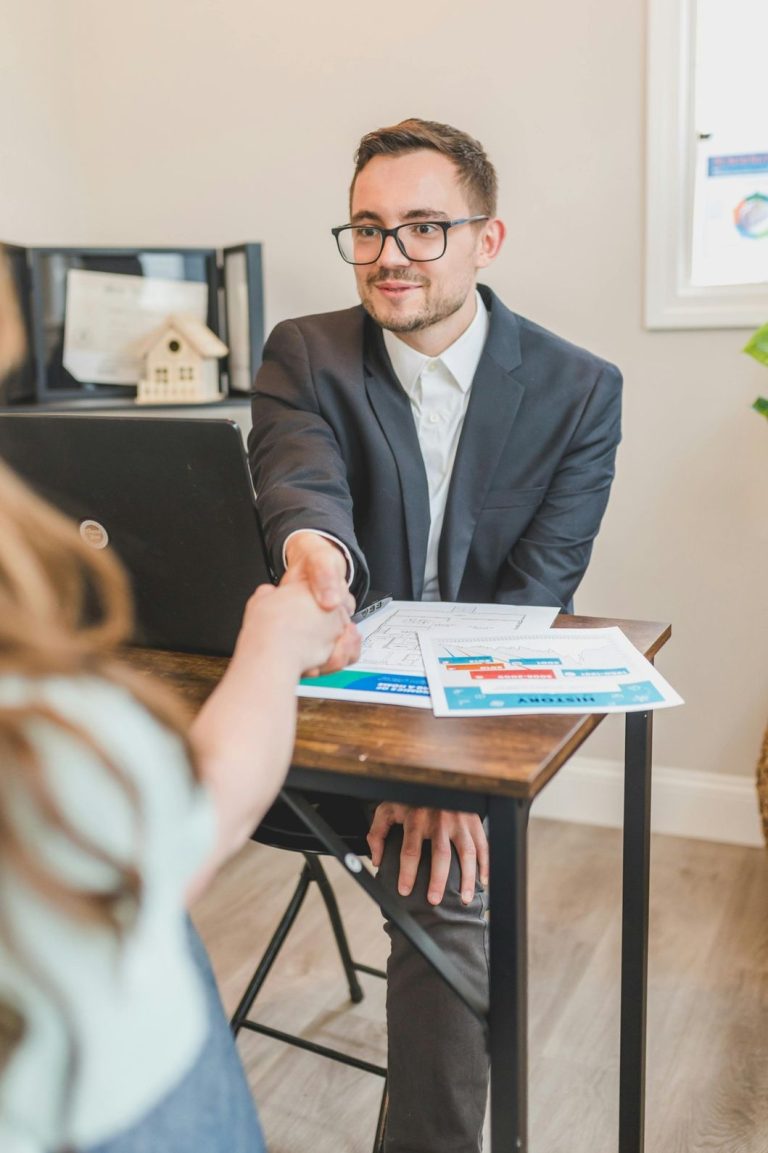 A realtor agent in Sugar Land shaking hands with a client at a desk with a laptop and real estate documents.