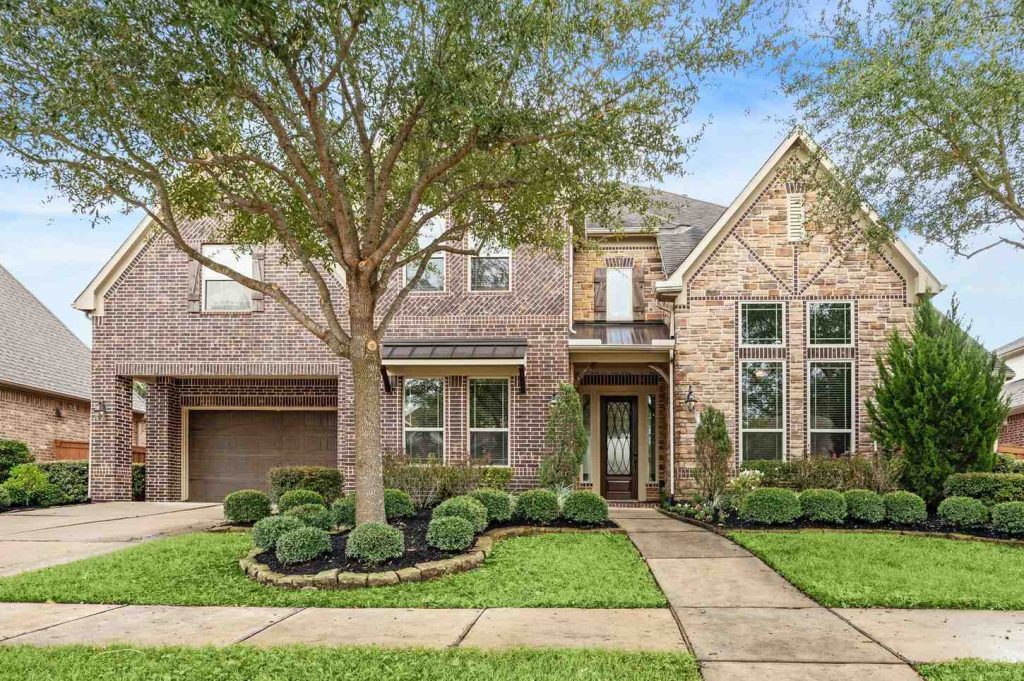 Spacious brick home with balcony and lush green backyard under blue sky, listed by Real Estate Brokers Sugar Land.