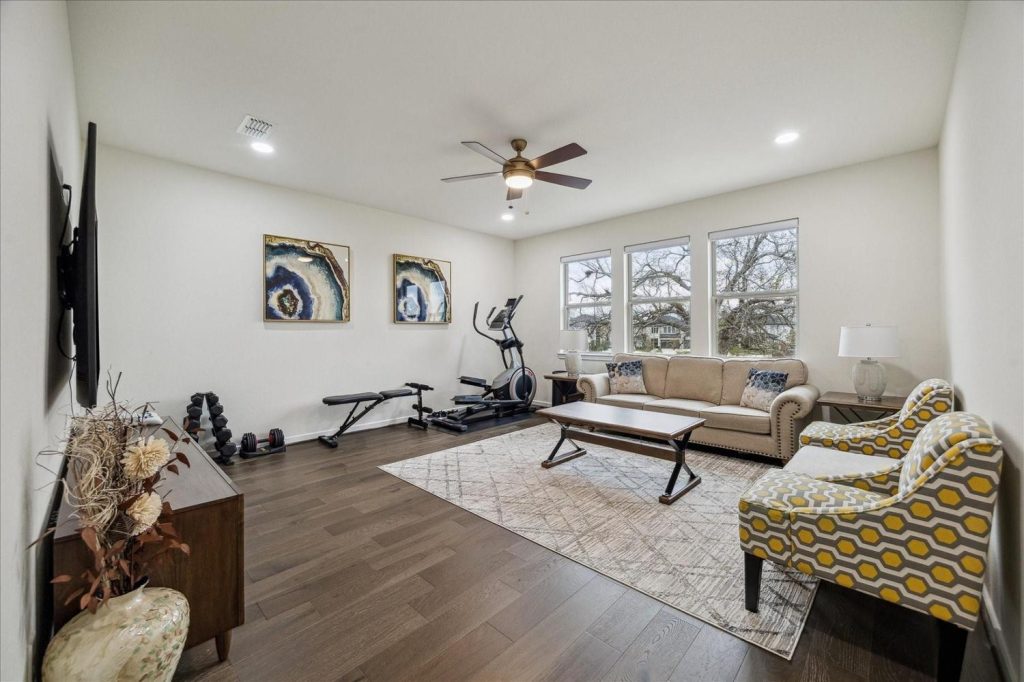 Bright home gym and lounge featuring elliptical, weights, cozy sofa, and large windows – Licensed Real Estate Agent Sugar Land
