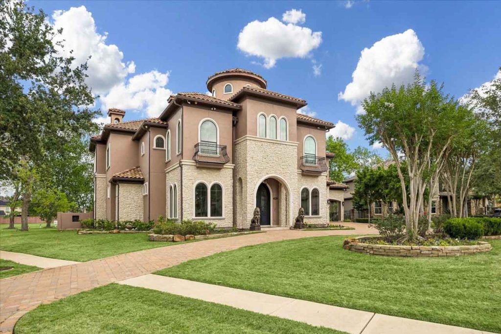Angled exterior view of a two-story Mediterranean home showing a brick walkway, arched windows, and manicured landscaping.