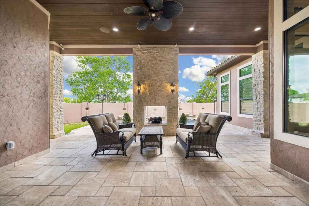 A covered outdoor patio featuring a large stone fireplace, tile flooring, and wicker furniture facing the open backyard.