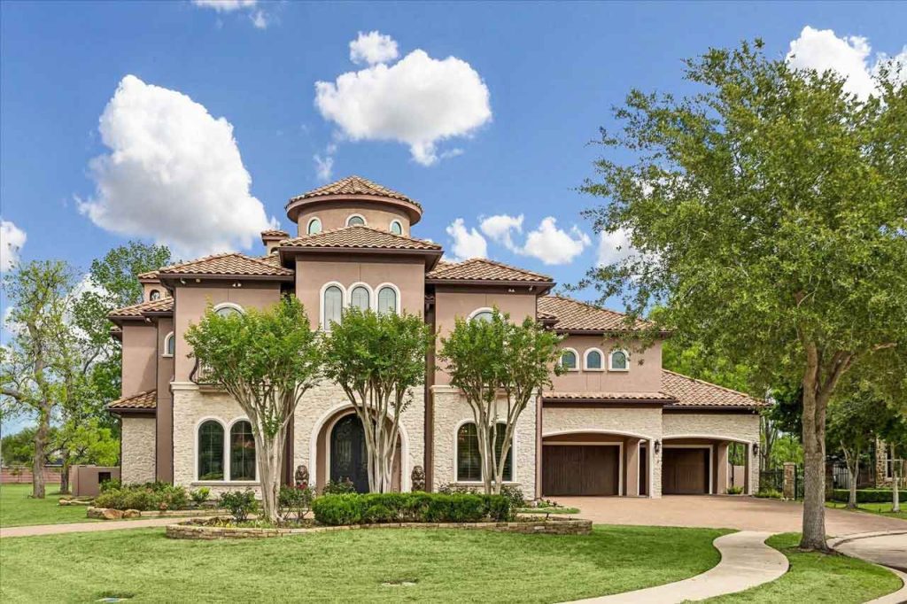Front view of a large Mediterranean-style house with stucco and stone walls, a tile roof, a turret, and a large green lawn.